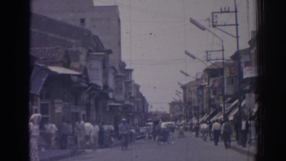 1962 Izmir street view with pedestrians moving past shop awnings, bicycles, streetlamps, and low-rise commercial buildings.