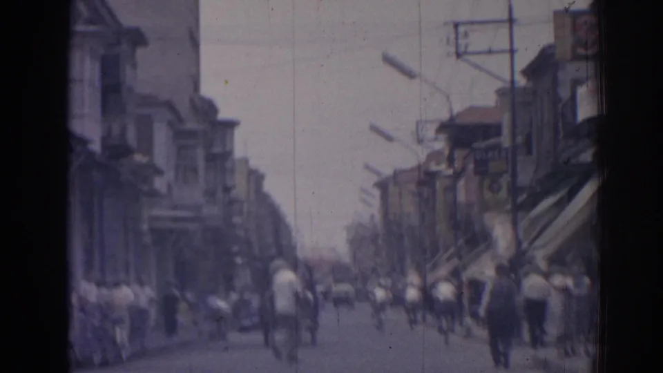 Soft 8mm view of a busy 1962 Izmir commercial street with pedestrians, bicycles, awnings, and storefront buildings.
