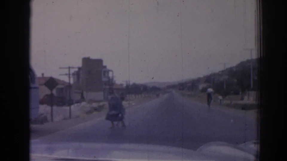 1962 Izmir road view from a moving vehicle, with pedestrians, roadside buildings, utility poles, and the vehicle hood at the bottom edge.