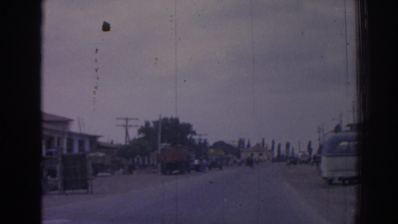 Roadway with pedestrians, trucks, utility poles, low buildings, and a bus-like vehicle at the right edge.