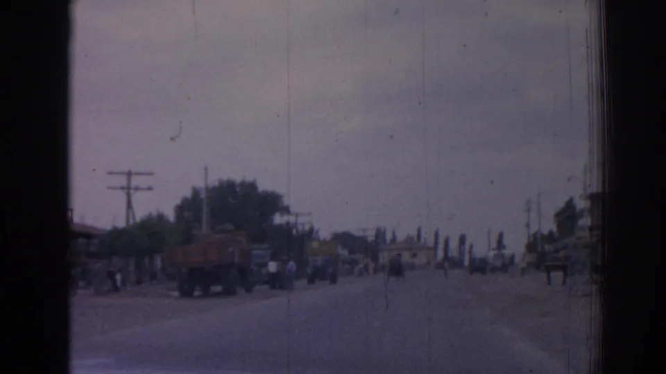 1962 Izmir street scene with a red truck, pedestrians, roadside trees, utility poles, and distant traffic under a hazy sky.