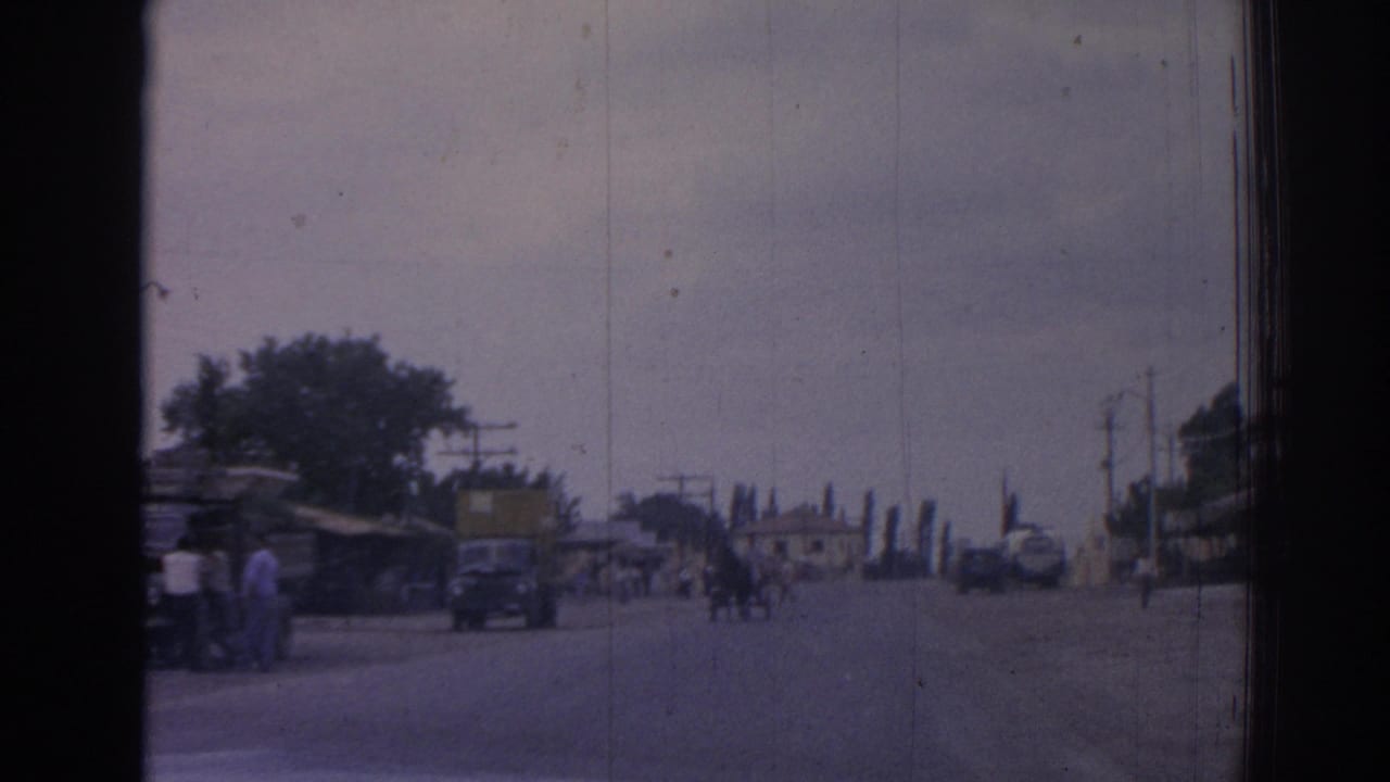 Street seen through dark frame edges with cars, pedestrians, trees, poles, and low buildings.