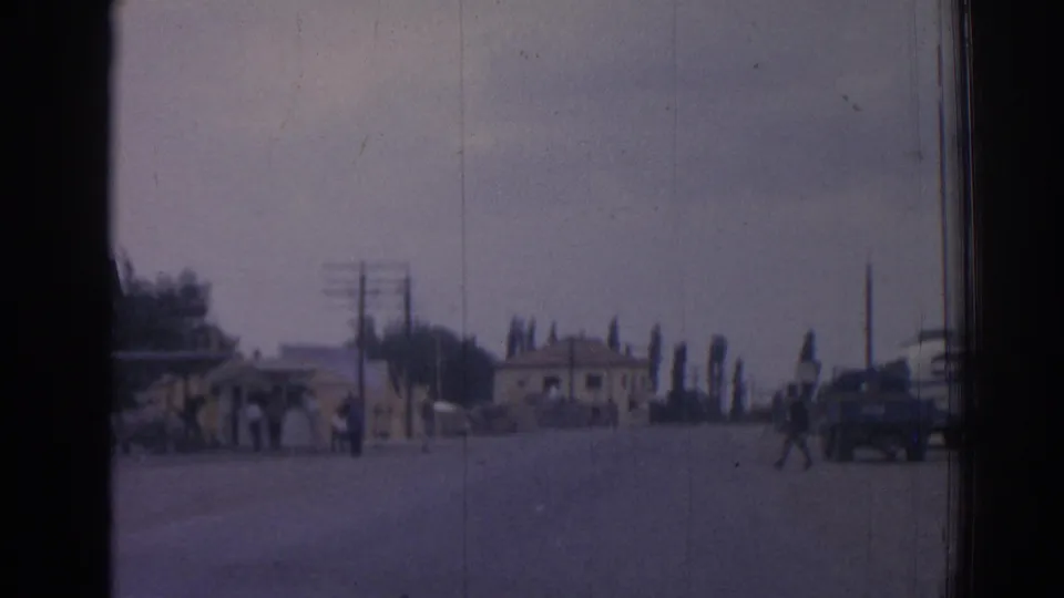 A broad 1962 Izmir roadway with pedestrians crossing, a dark mid-century car at right, utility poles, trees, and low roadside buildings.