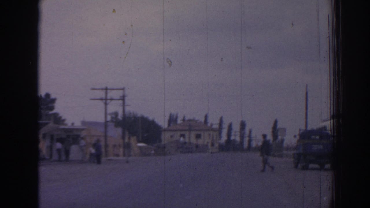 Paved road with utility poles, small roadside building, pedestrians, and a parked vehicle in the distance.