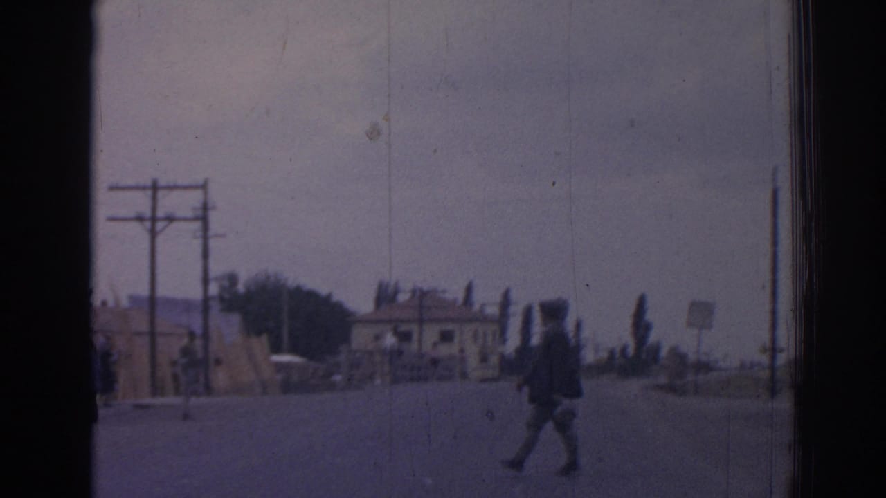 Man crossing a wide road near utility poles, a small building, roadside barriers, and a dark parked truck.