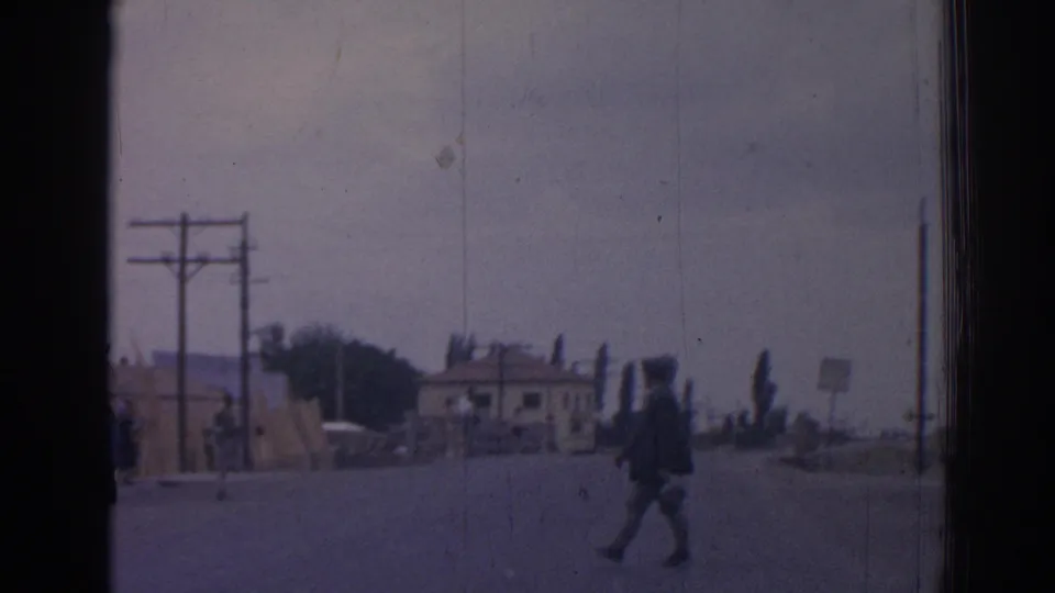 A pedestrian crosses a wide road in 1962 Izmir near utility poles, roadside barriers, a small building, and a parked truck.