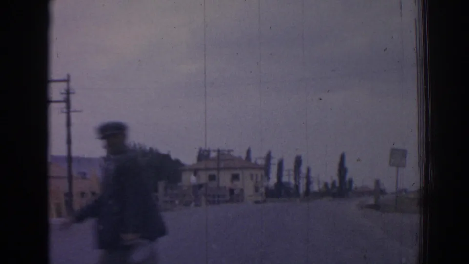 A blurred uniformed-looking figure passes near the road in 1962 Izmir, with utility poles, a small building, and a roadside sign behind.