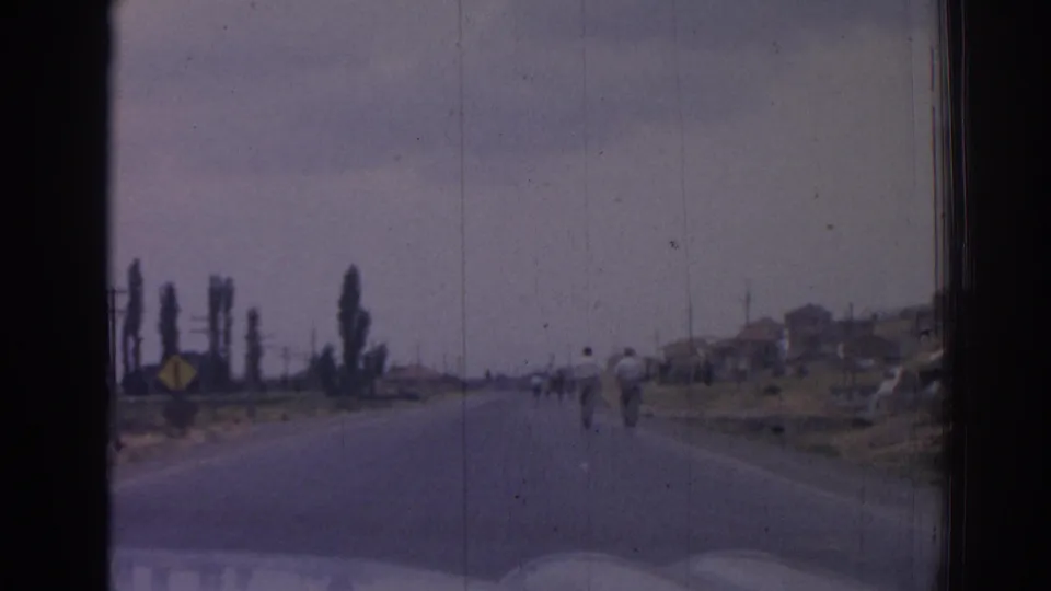 The road continues ahead in 1962 Izmir, with distant pedestrians, utility poles, roadside signs, trees, and low buildings along the horizon.