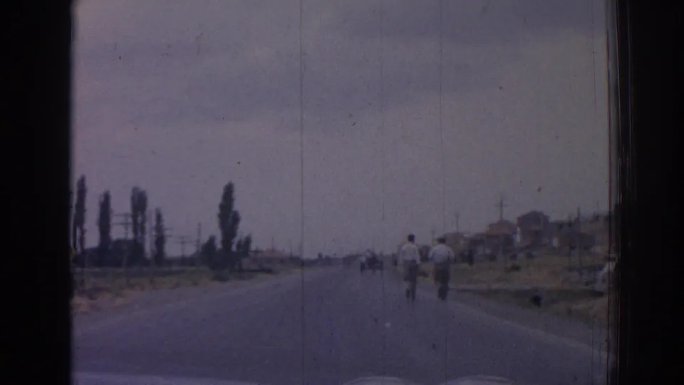 1962 Izmir home-movie frame from a moving vehicle, showing two men walking away on a paved road with poplars and roadside buildings.