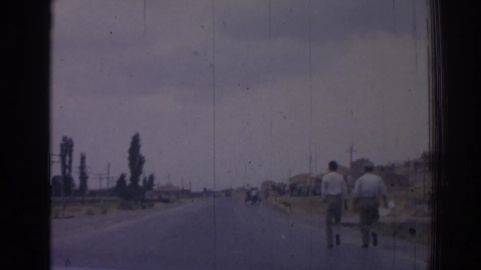 1962 Izmir road view with two men walking away at right, distant pedestrians or riders ahead, trees, utility poles, and low roadside structures.