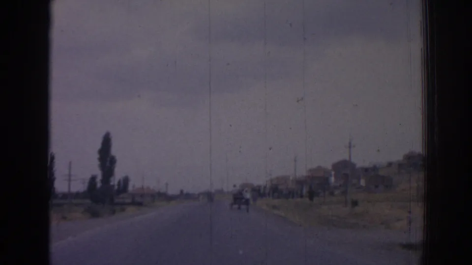 1962 Izmir home-movie road view, with a small horse-drawn cart ahead, sparse buildings to the right, trees to the left, and visible film wear.