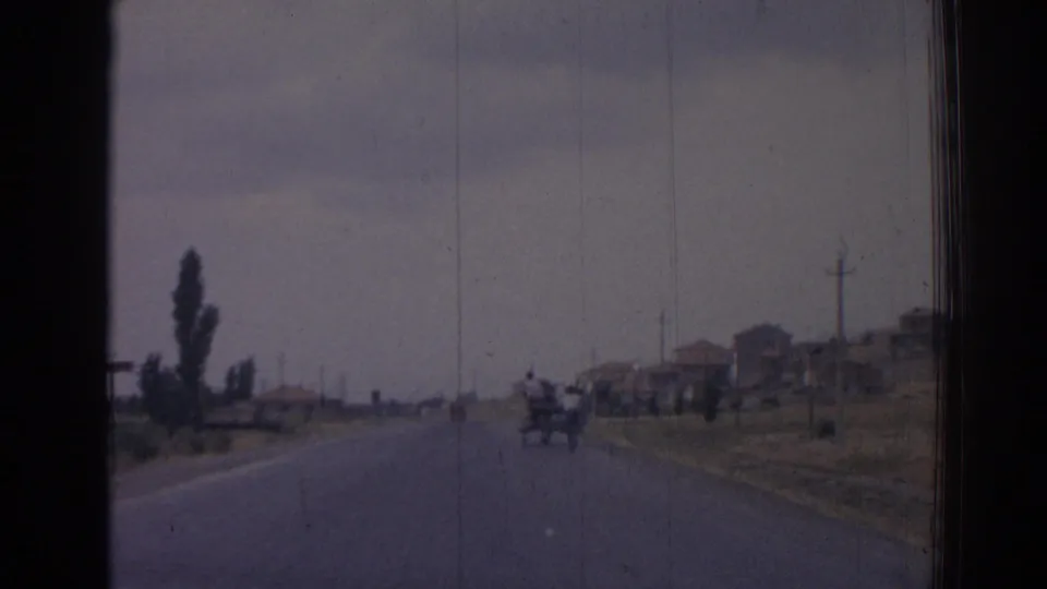 1962 Izmir road scene from a moving vehicle, with distant figures or cart traffic ahead, poplar trees, utility poles, and modest buildings under a cloudy sky.
