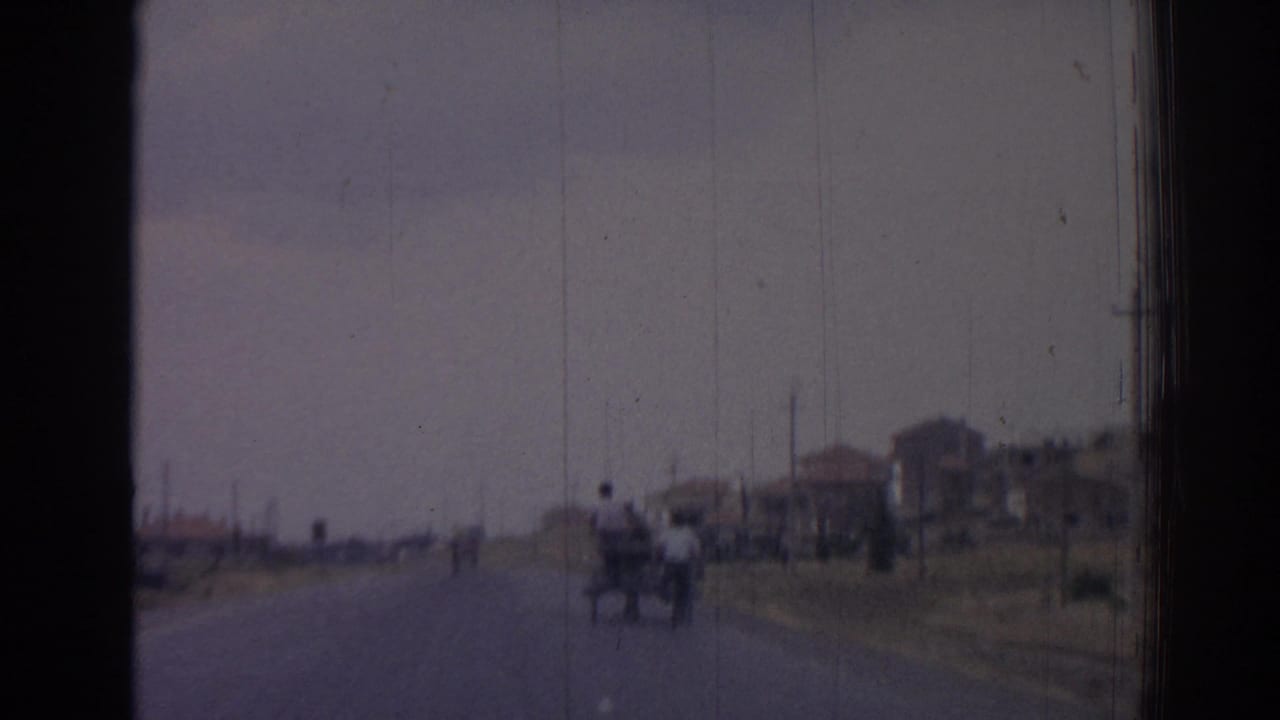 1962 Izmir home-movie frame showing a rural road, distant cart, modest roadside houses, and utility poles through a scratched 8mm image.