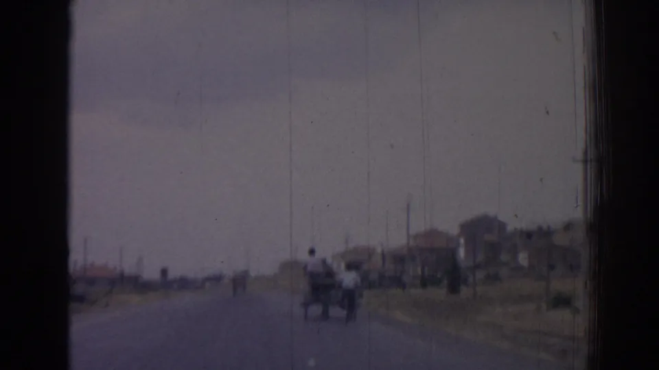 1962 Izmir home-movie frame showing a rural road, distant cart, modest roadside houses, and utility poles through a scratched 8mm image.