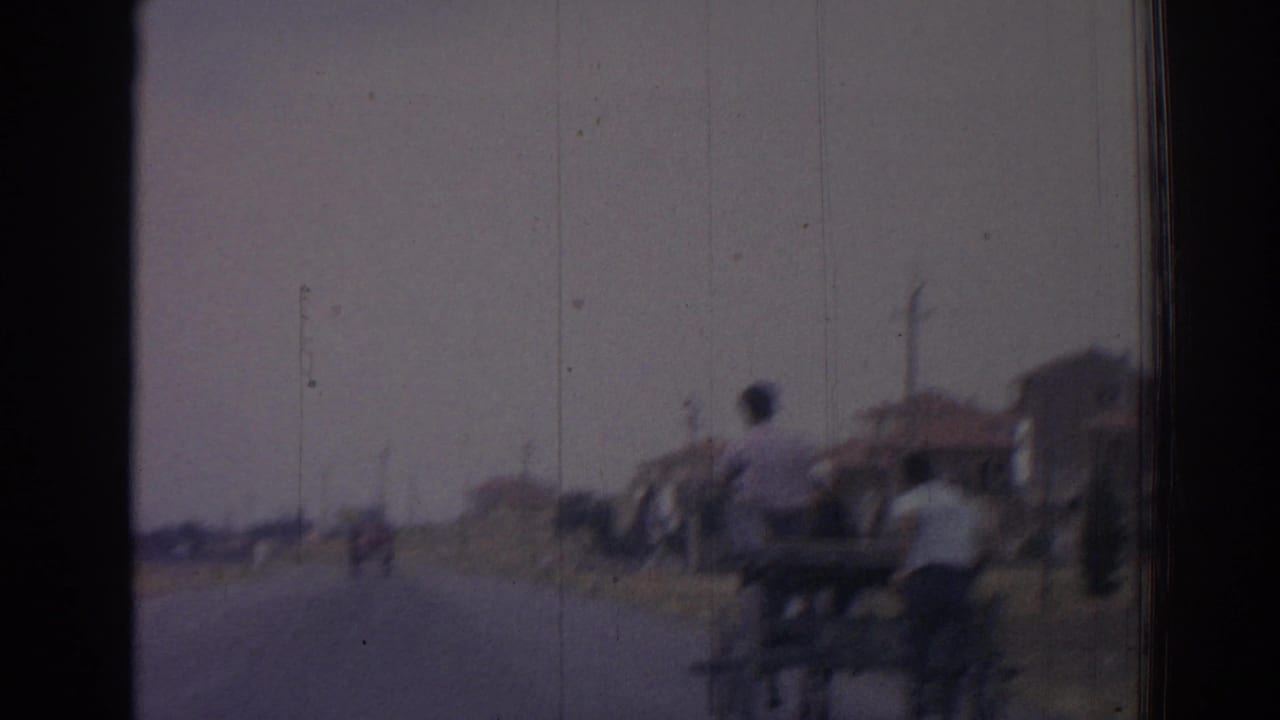 A man with a two-wheeled cart appears beside the road as houses recede in the background.