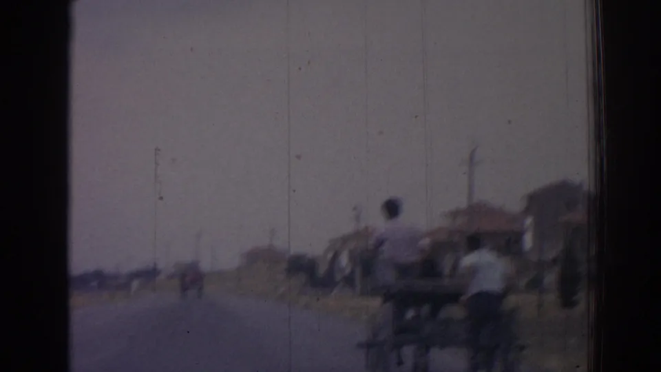 In this 1962 Izmir roadside view, a man and two-wheeled cart pass near the camera with low houses and a hazy road behind.