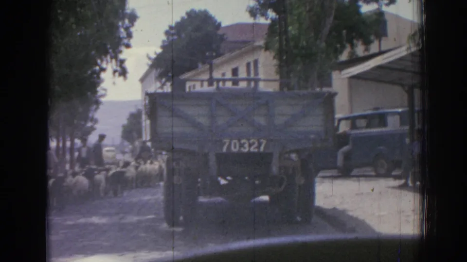 1962 Izmir home-movie frame showing sheep on a shaded town street near a canvas-covered truck marked 70327 and low buildings.