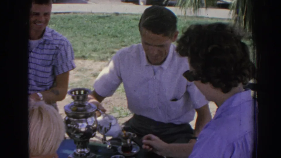 1962 Izmir home-movie frame showing adults seated outdoors around a shiny metal samovar-like vessel, with grass and a tree nearby.