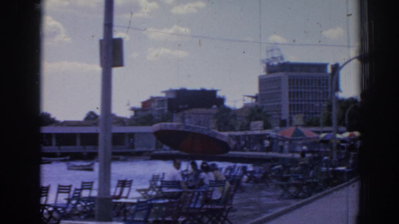 Harbor promenade with empty chairs, seated people, boats, buildings, and a red umbrella.