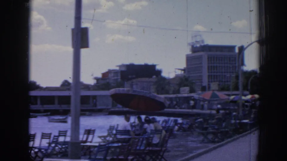 1962 Izmir harbor view with empty promenade chairs, a few seated people, small boats, waterfront buildings, and a red umbrella.