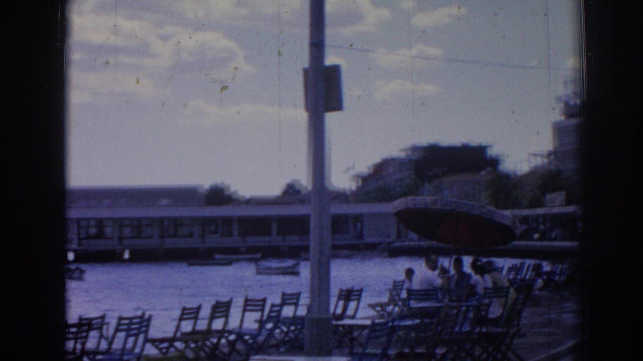 Waterfront chairs beside harbor water with a large boat, pier building, people, and pole.