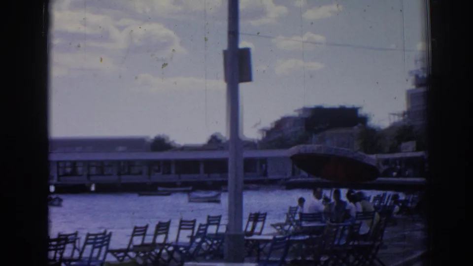 1962 Izmir waterfront scene showing rows of chairs, a few people, harbor water, a large boat, pier structure, and nearby buildings.