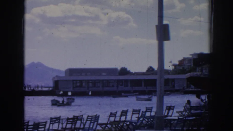 1962 Izmir harbor view with empty promenade chairs, small boats on the water, a long pier building, a pole, and mountains in the distance.
