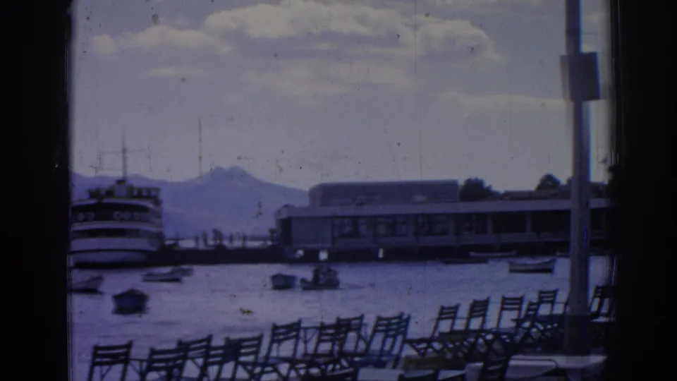 1962 Izmir harbor scene with rows of chairs, small boats, a larger vessel, long waterfront structure, pole, clouds, and distant mountains.