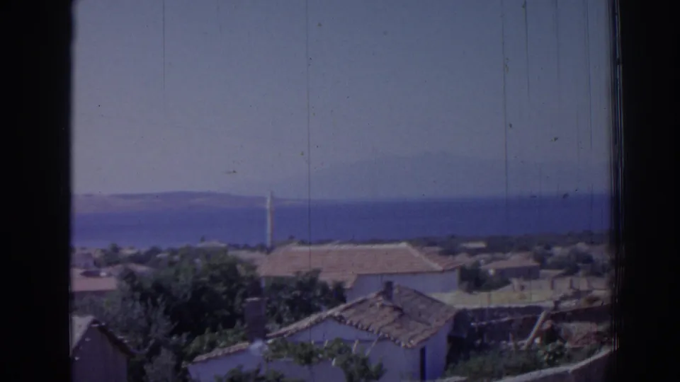 1962 Izmir 8mm still with foreground rooftops and trees, a blue coastal horizon, distant hills, and a narrow minaret.