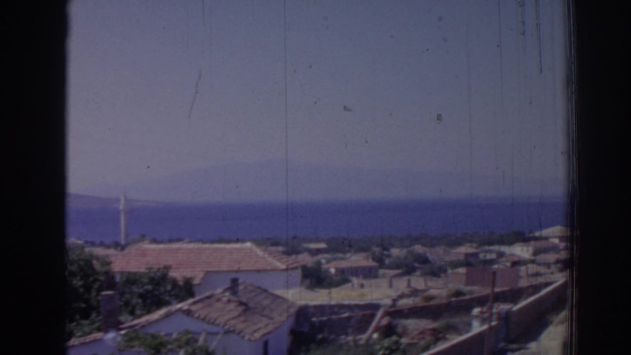 1962 Izmir coastal village view with terracotta roofs, blue sea, hazy sky, and a dark vertical window frame.