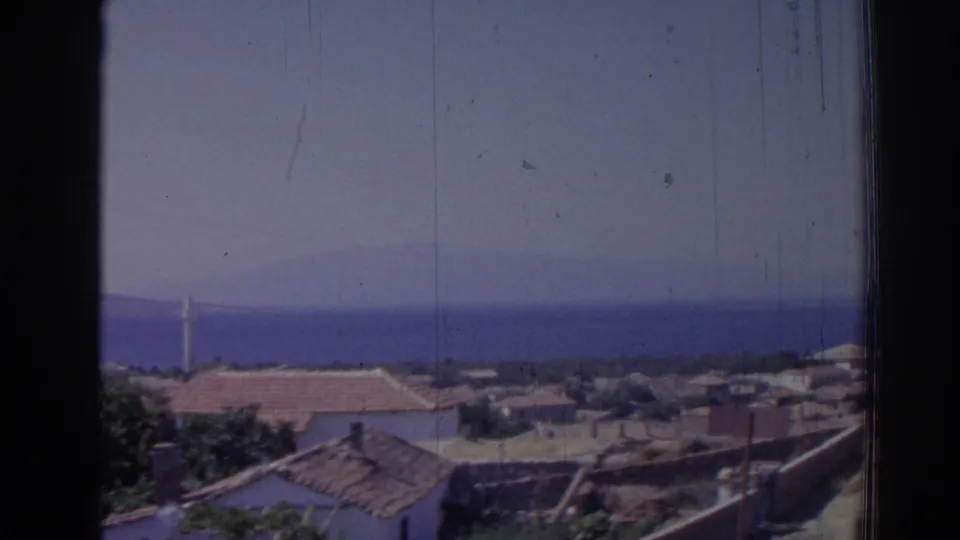 1962 Izmir coastal village view with terracotta roofs, blue sea, hazy sky, and a dark vertical window frame.