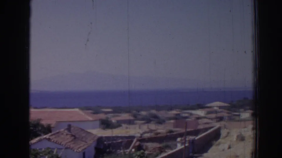 1962 Izmir shoreline scene with low houses, terracotta roofs, open pale ground, blue water, and dark window framing.