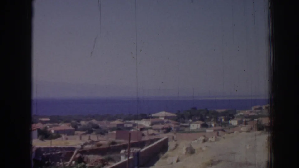 1962 Izmir home-movie frame with coastal houses, pale hillside ground, blue sea, and a dark window edge.