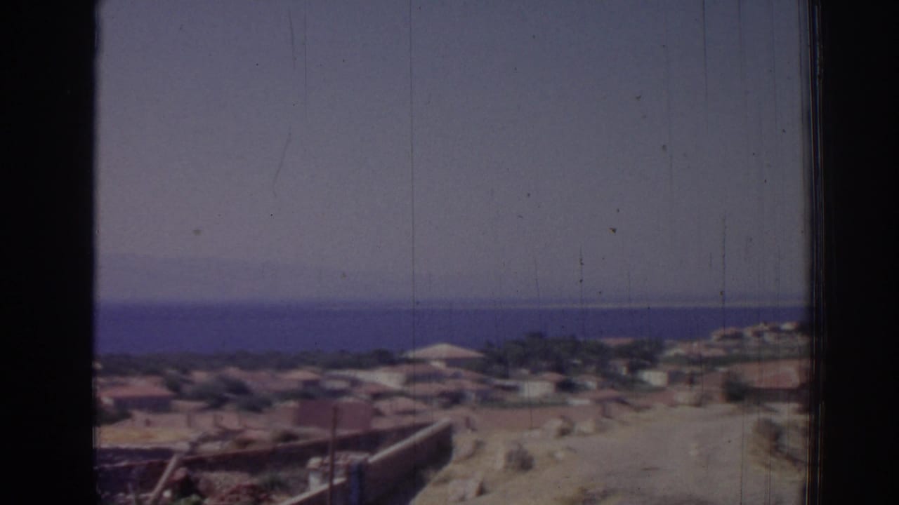 Elevated view of a coastal town with low buildings, sandy hillside, and blue sea beyond.