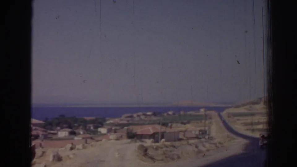 1962 8mm Izmir coastal panorama with a hillside road, low buildings, sandy slopes, and distant sea.