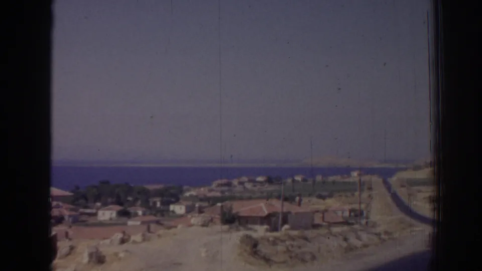 1962 Izmir-area coastal village seen from a road, with low houses, dry ground, utility poles, and blue sea beyond.