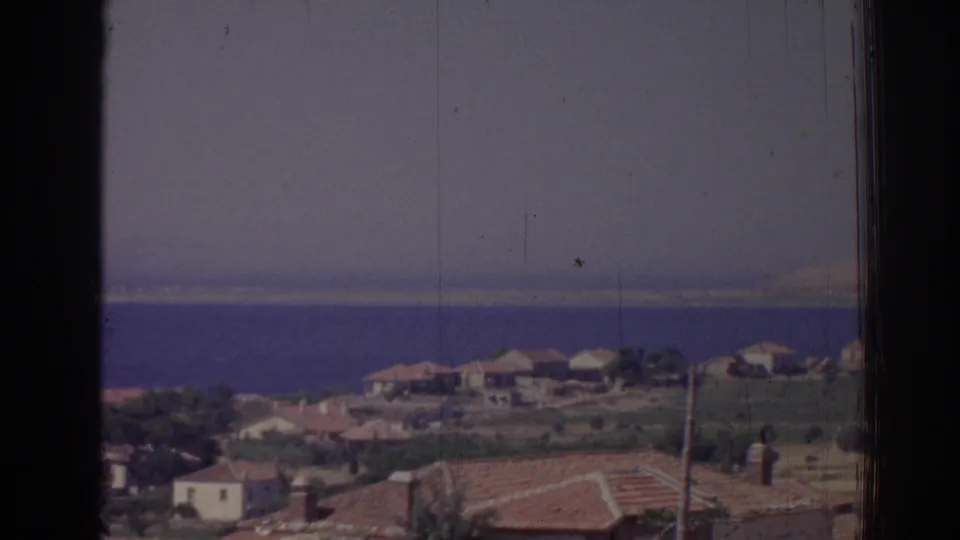 A closer road-view frame of Izmir coastal houses, rooftops, dry ground, and blue sea on faded 8mm film.