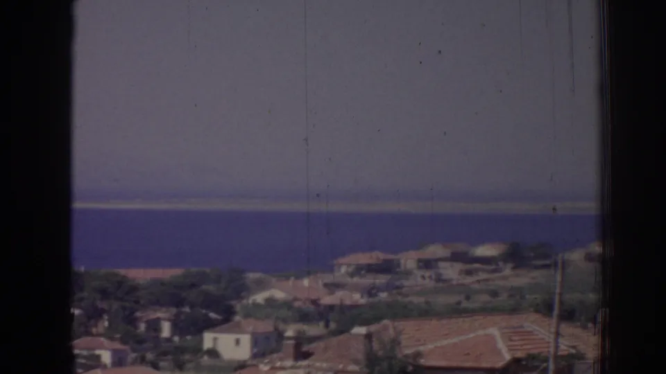 Izmir coastal village rooftops and scattered buildings appear beneath a hazy sky with the Aegean-looking sea beyond.