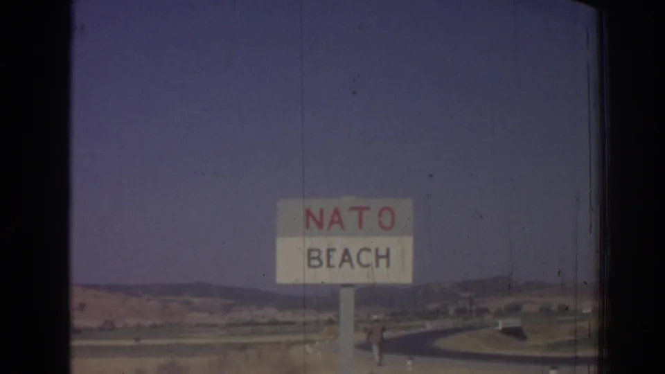 1962 8mm frame from Izmir, Turkey, showing a roadside NATO Beach sign with hills and a hazy sky behind it.