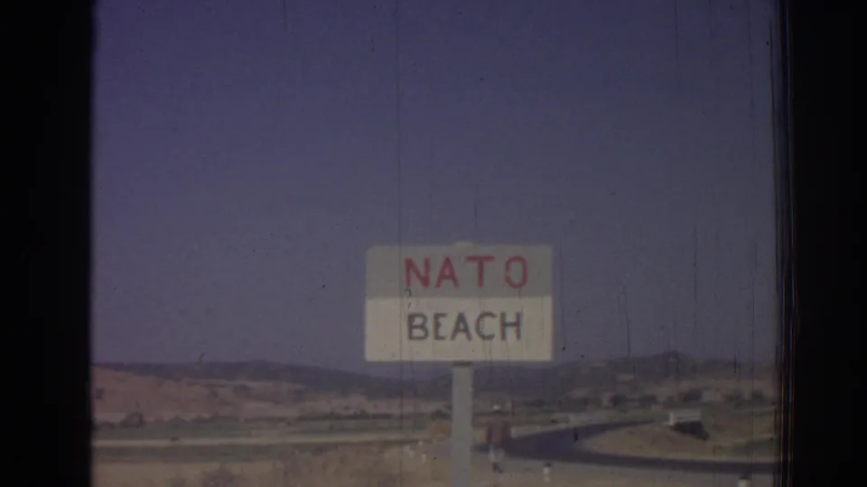 Roadside NATO Beach sign in a 1962 Izmir home-movie frame, with a road, low hills, and hazy sky visible.