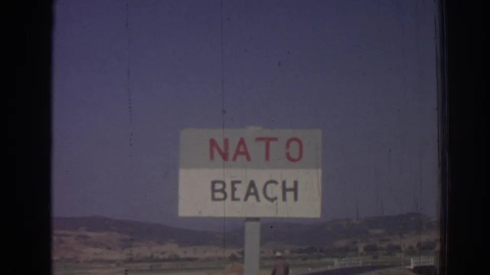 Closer 1962 8mm view of the NATO Beach sign near Izmir, with distant hills, road edge, and faded film color.