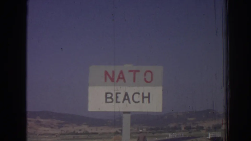 Final 1962 Izmir frame showing the NATO Beach sign centered against a hazy hillside landscape and roadside foreground.