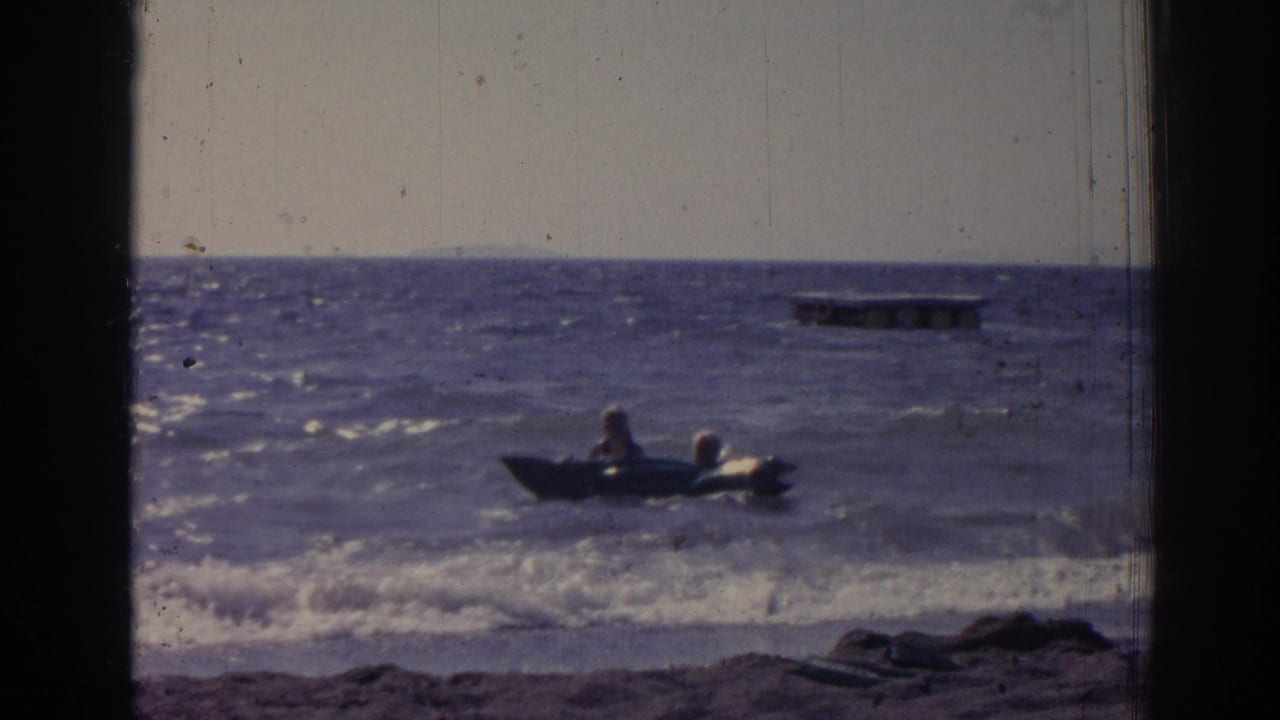 Two people sit in a small raft in choppy water near shore.