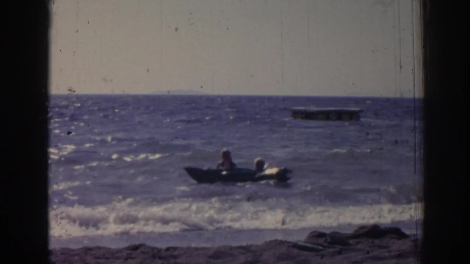 Two bathers ride a small dark inflatable raft in the surf off an Izmir beach, with a floating platform in the distance.