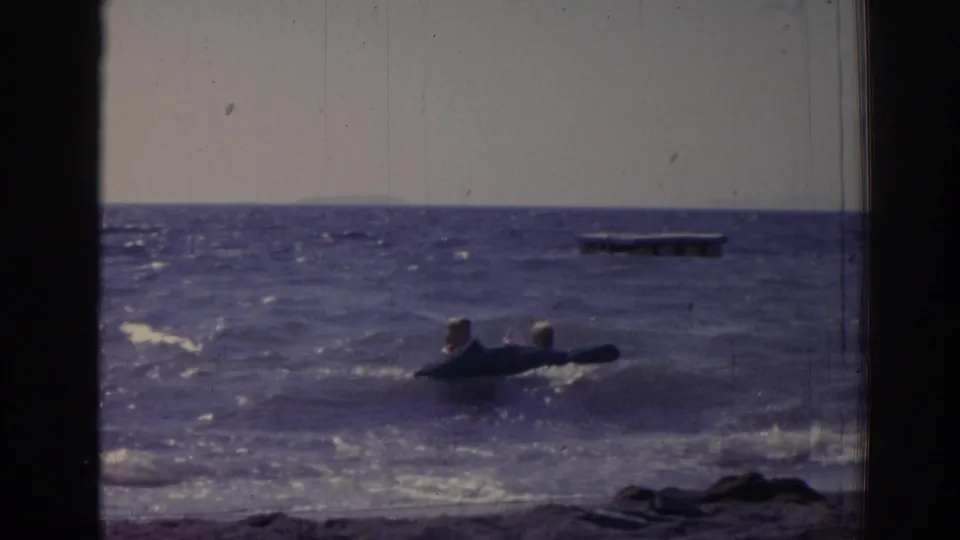 The raft drifts through shallow surf while a distant floating platform sits beyond it on the open water.