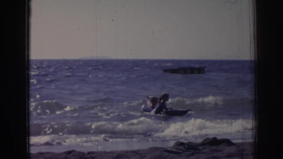 Two people sit upright in a small raft as a wave breaks near shore, filmed in 1962 home-movie footage from Izmir.
