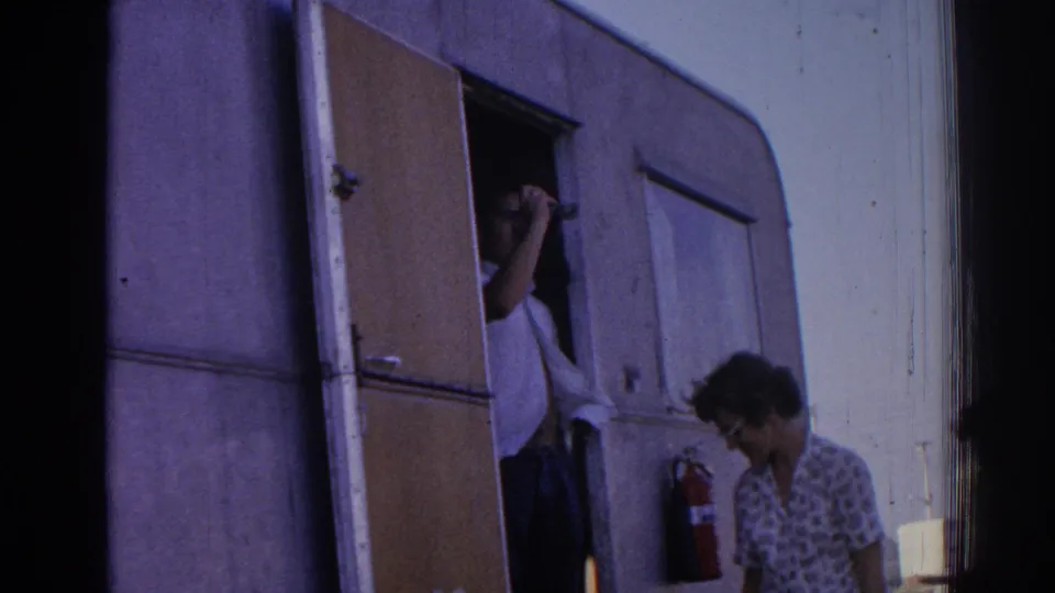 A woman in patterned clothing stands near a trailer doorway in 1962 Izmir footage as a person appears partly inside.