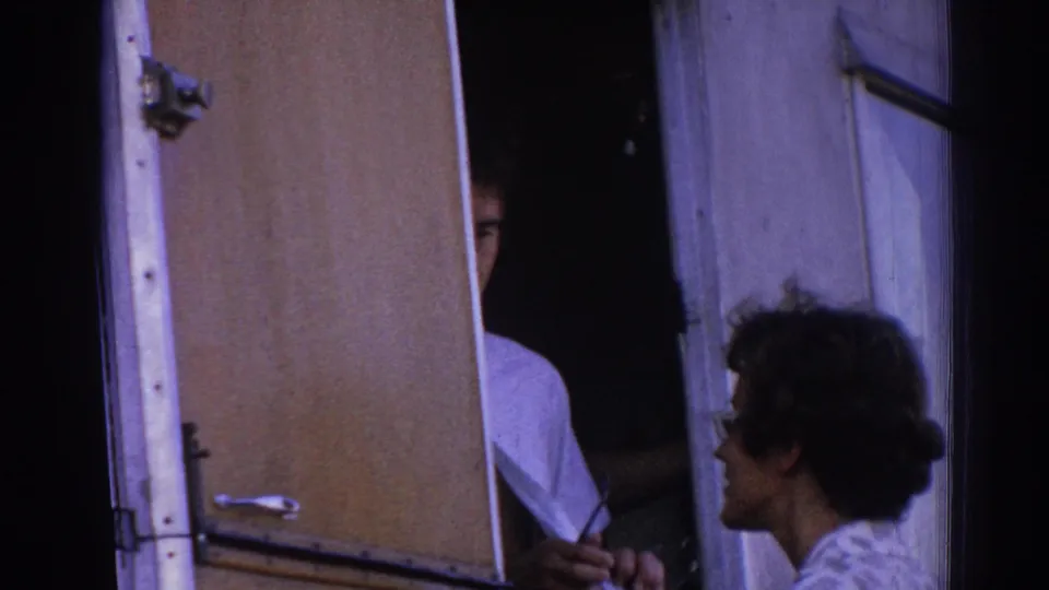 A close handheld view shows a man partly hidden in the trailer doorway while a woman in sunglasses looks toward him in Izmir.
