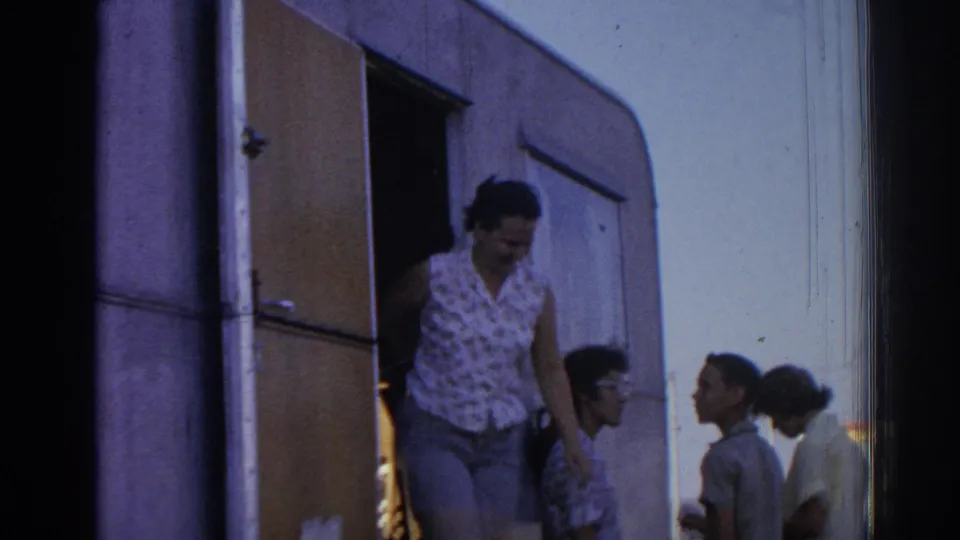 1962 Izmir campground frame with a woman leaving a camper trailer as adults gather beside it.