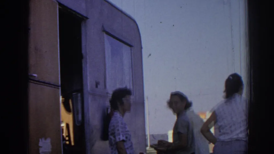 1962 Izmir home-movie frame showing adults beside a camper, including a woman in sunglasses and another with a ponytail.